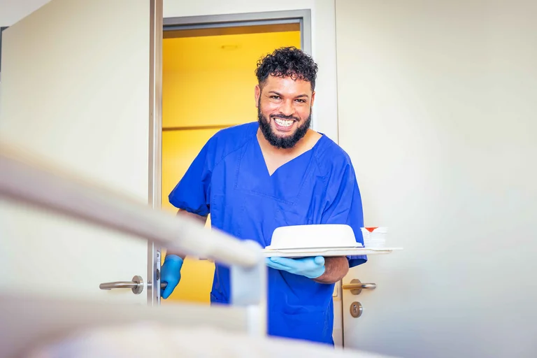 Ein Pflegefachassistent bringt lächelnd ein Tablett mit Essen in ein Patientenzimmer.