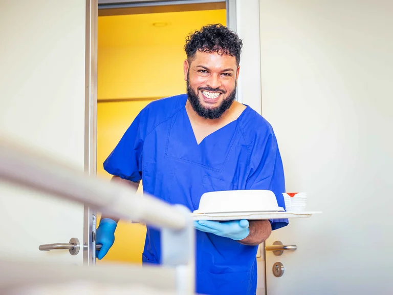 Ausbildung Pflegefachassistenz: ein Auszubildender bringt lächelnd ein Tablett in eine Patientenzimmer