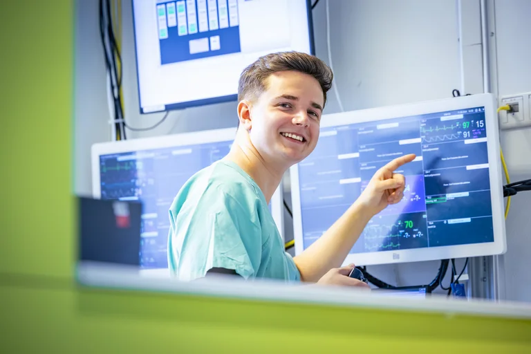 Der ehemalige Praktikant steht im Rahmen seines Praktikums am Stützpunkt der Pflegefachkräfte auf einer Station im Krankenhaus und zeigt lächelnd auf einen Bildschirm.