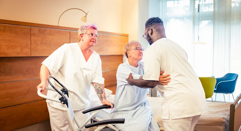 Das Bild zeigt zwei Auszubildende, die einer älteren Patientin aus dem Bett in einen Rollstuhl helfen.  