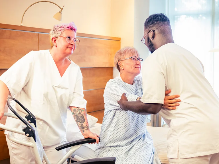 Ausbildung Pflegefachassistenz: Zwei Auszubildende helfen einer Patientin aus dem Bett in einen Rollstuhl